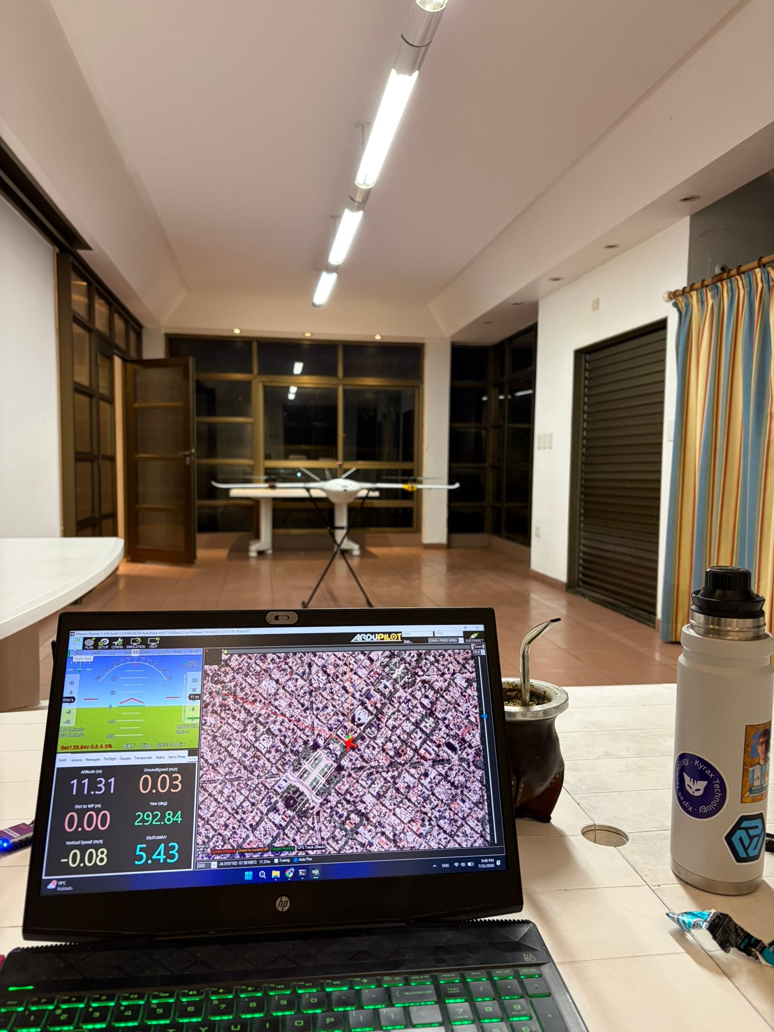 La estación de tierra: una notebook con el software de vuelo y el mapa satelital, y el drone al fondo sobre su soporte, en un local.
